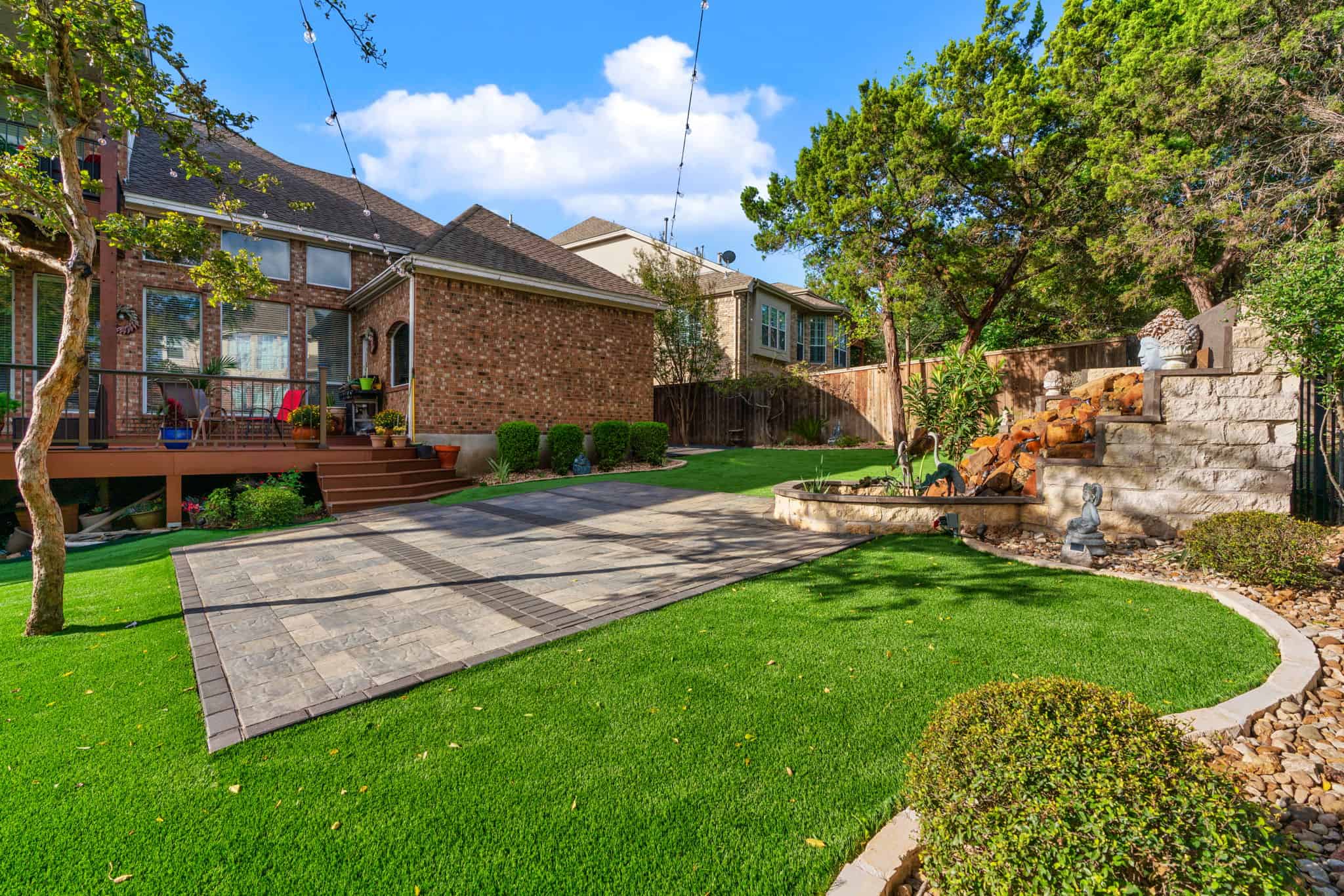 a large landscaped backyard with a brick house, wooden deck, and artificial turf for pets