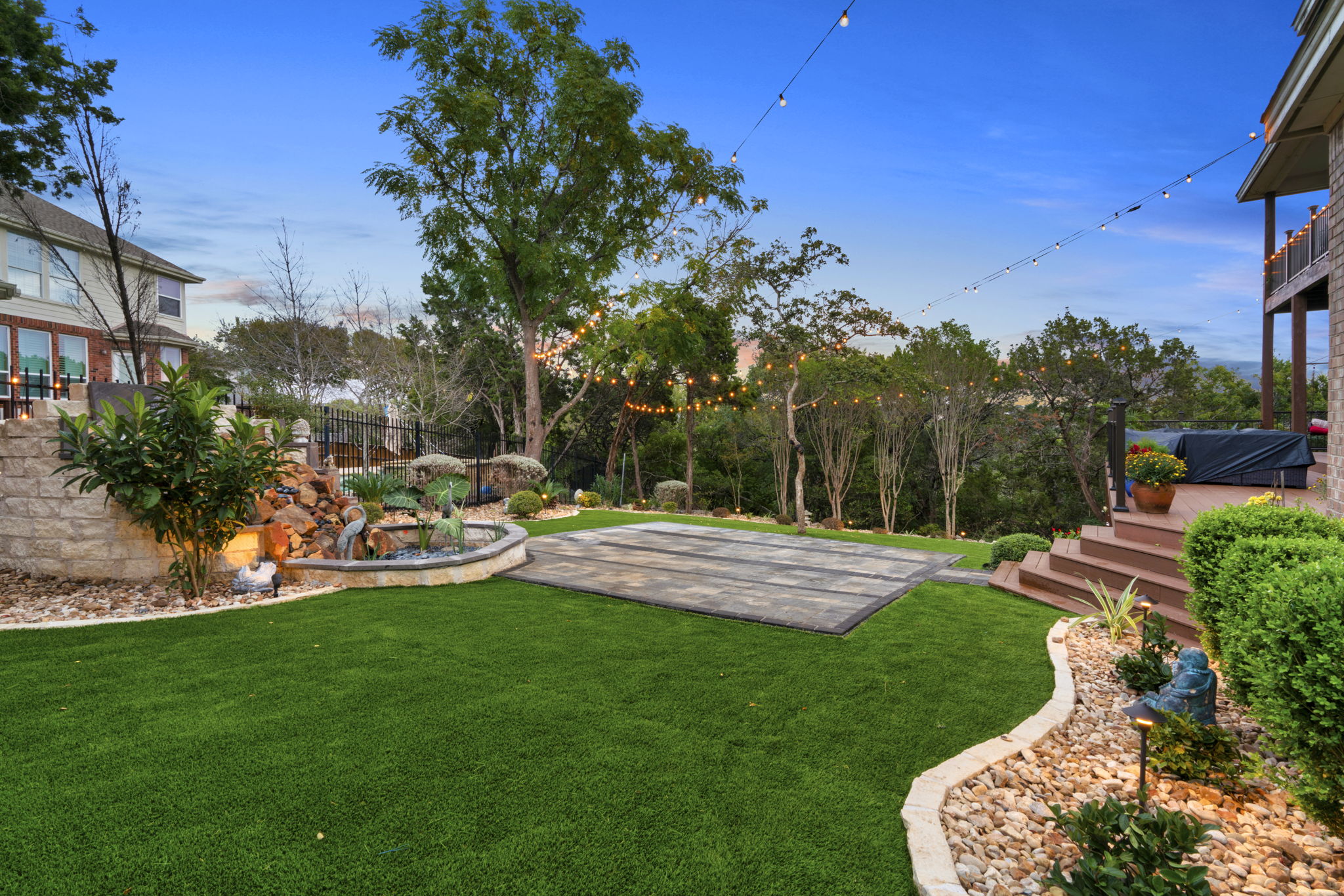 a large stone patio outside a wrought iron fence with a zen fountain and trees with hanging lights