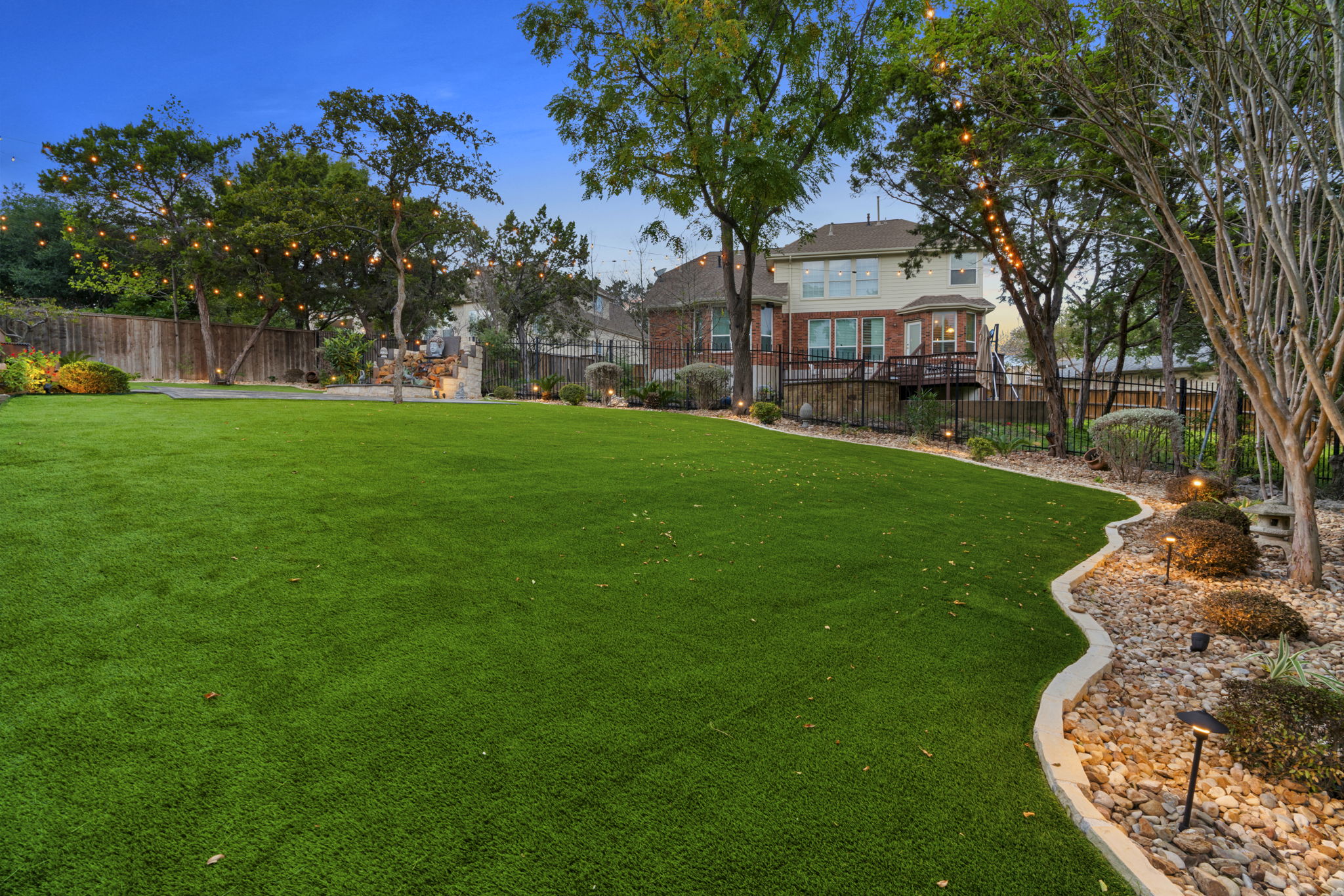 a large house with an artificial turn lawn surrounded by a wrought-iron fence and landscaped mulch beds with trees