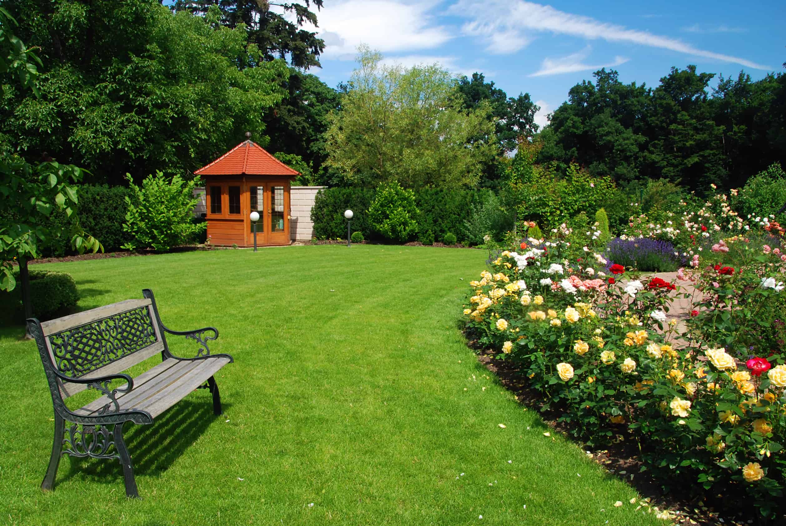 a green lawn with summer flowers next to a wood bench with a covered pergola in the background