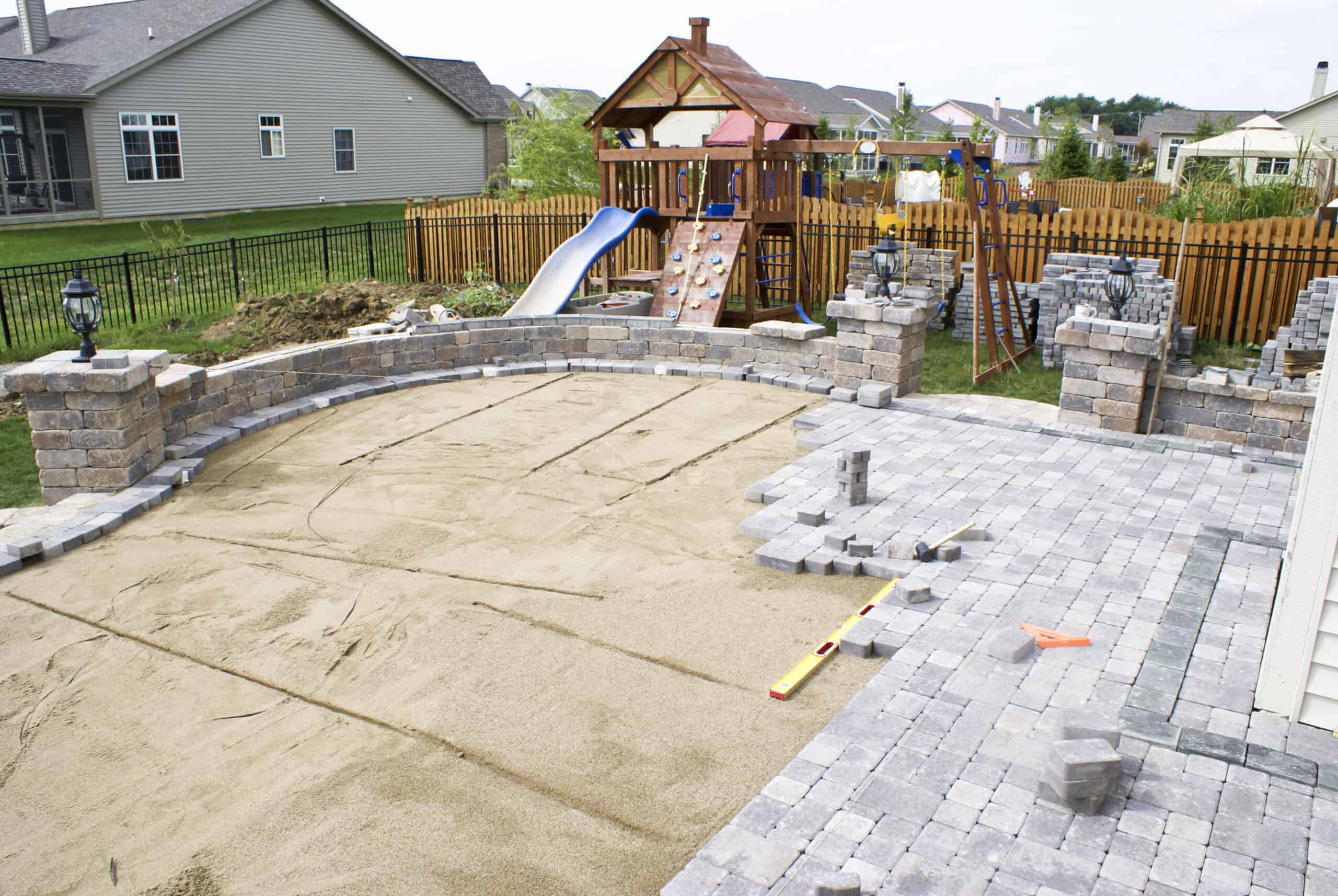 a large paver patio with retaining walls under construction in a fenced-in backyard with a playground