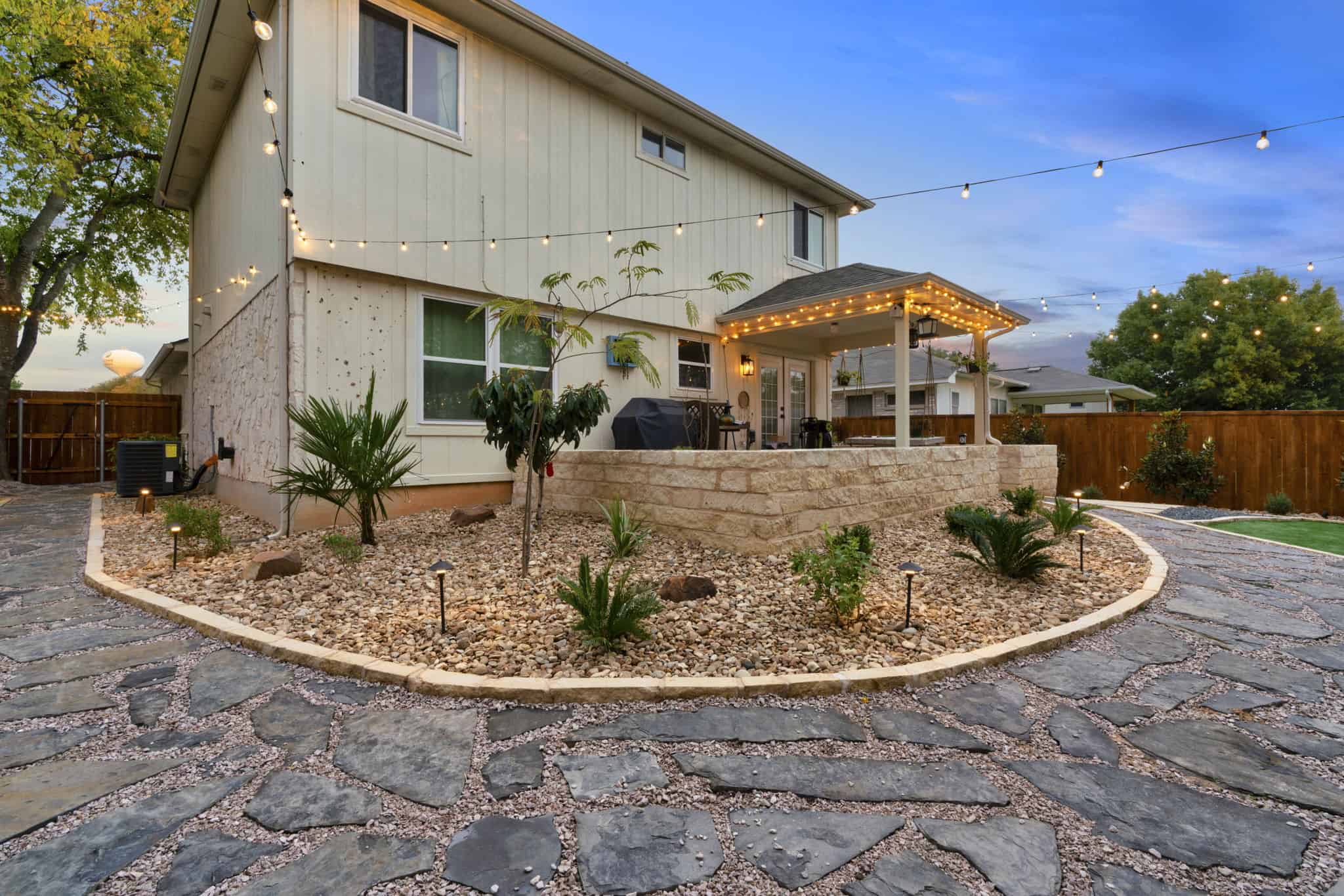 Two-story home with pergola-covered patio, stone walls, string lights, gravel landscaping, and curved stone pathway.