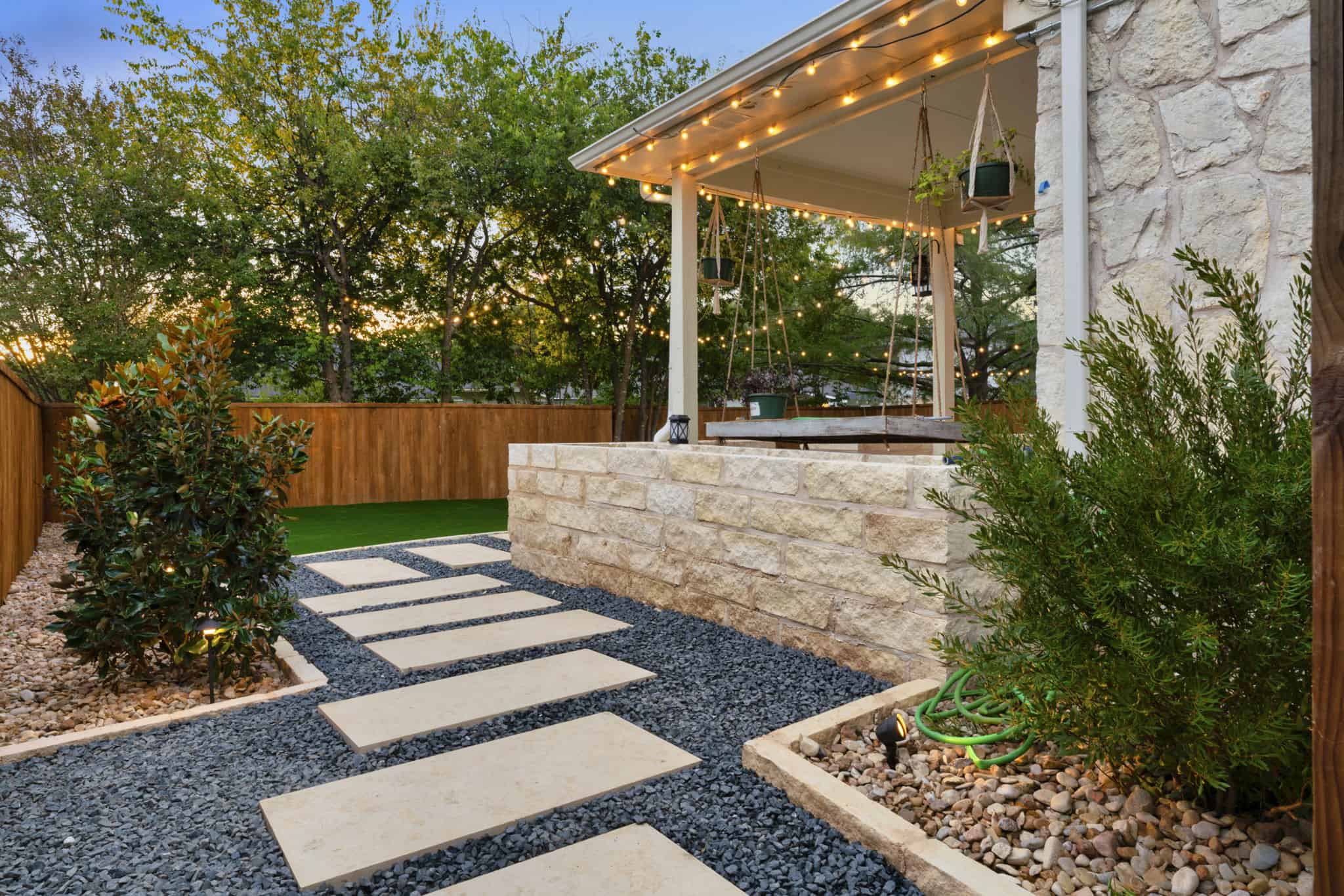 Modern backyard patio with pergola, stone wall, string lights, hanging plants, and paver path surrounded by trees.