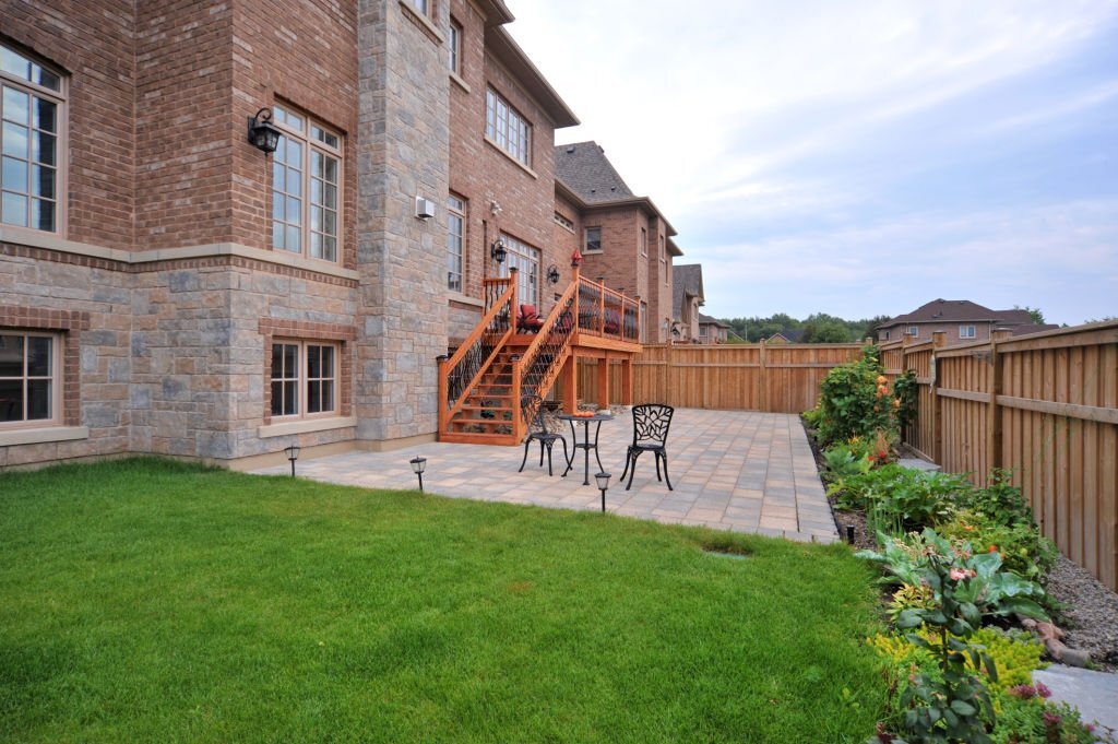Stone patio with wrought iron furniture, wooden stairs, and garden beds alongside a large brick house with lush green lawn.