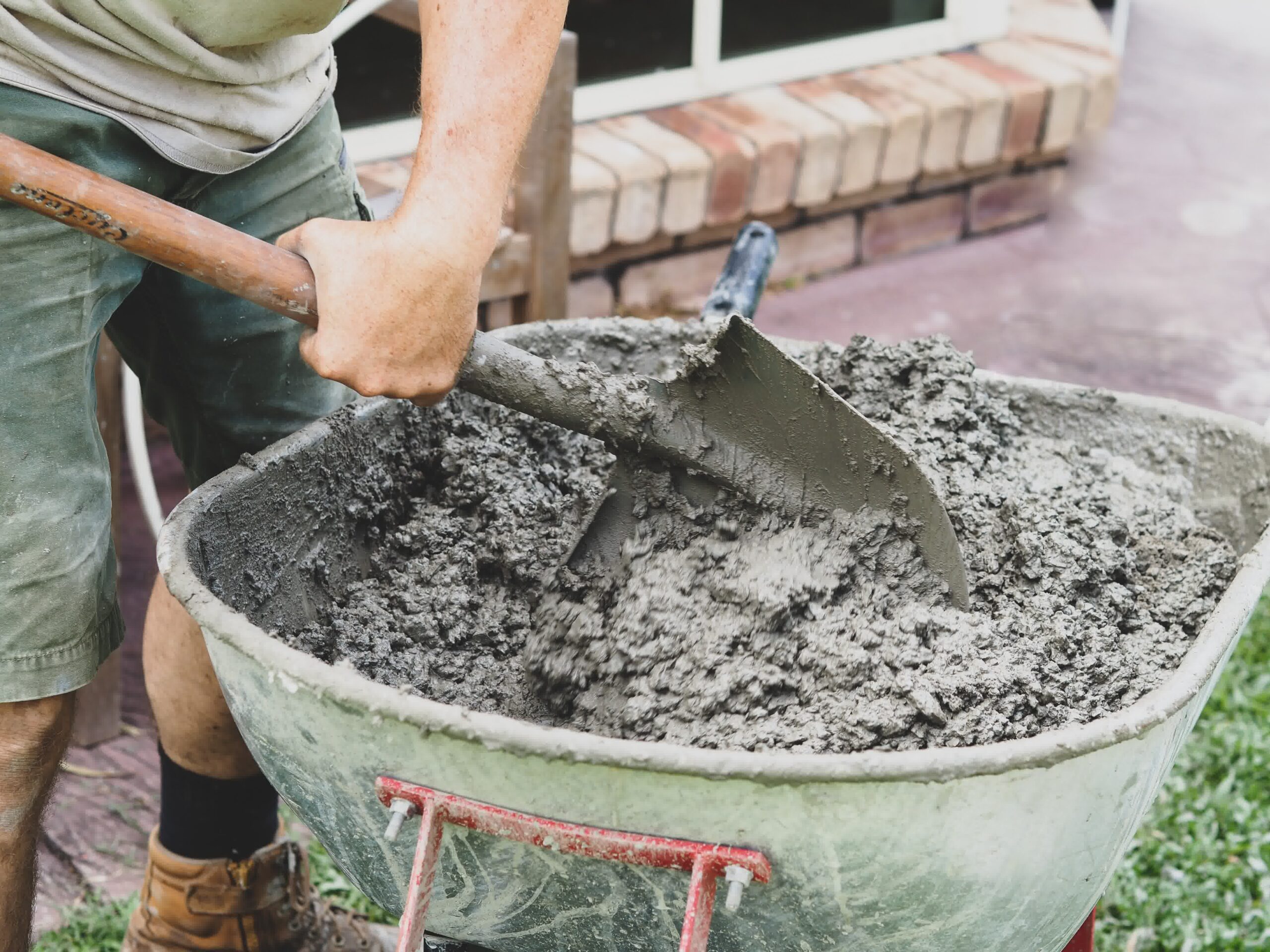 Worker mixes wet concrete in a wheelbarrow using a shovel during residential construction, preparing for sidewalk or patio pour.