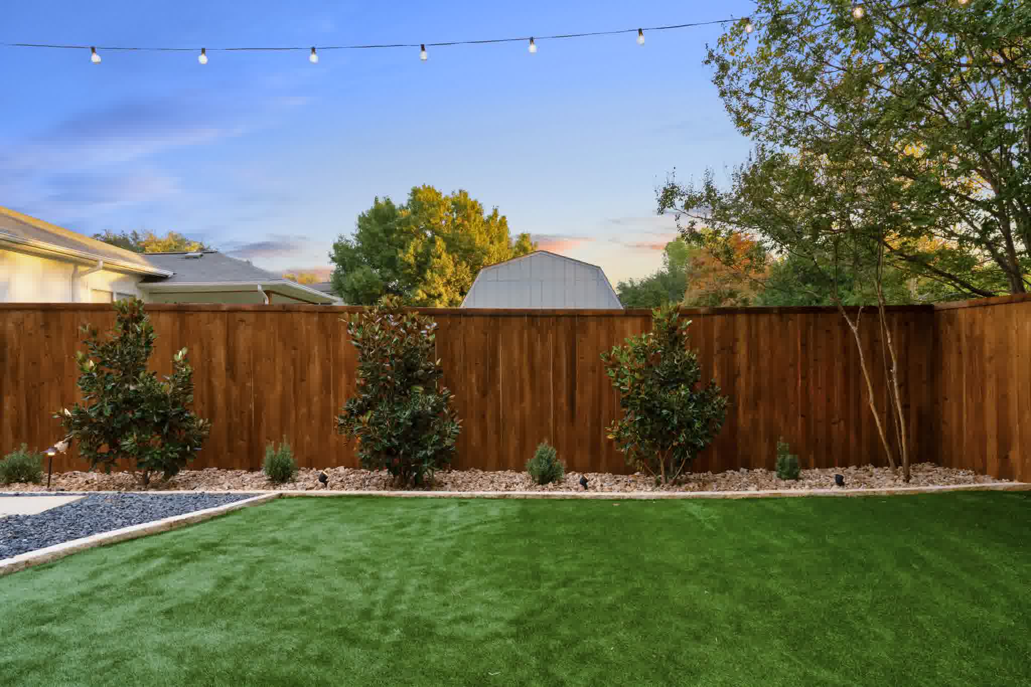 Tall wooden privacy fence with landscaped garden beds and string lights framing a lush green lawn at sunset.